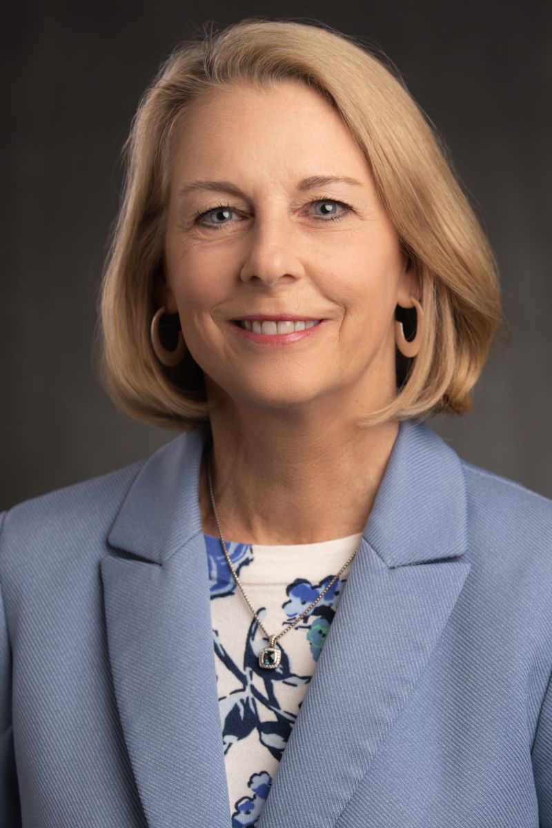Tammy Shepherd, a Caucasian female with short blonde hair wearing gold earrings,a silver necklace with a blue stone, a white shirt with blue patterns, and a blue suit jacket.
