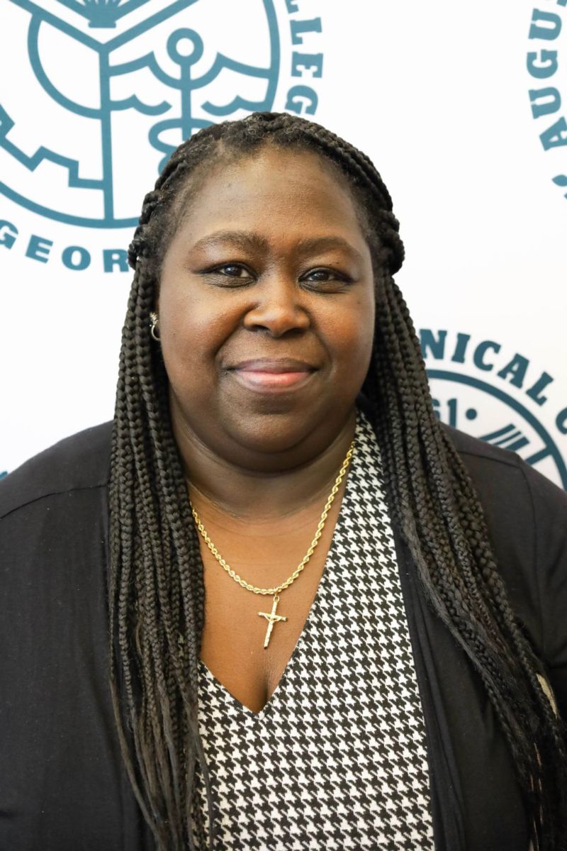 Tiffany Rowe-Thomas, an African American female with long braided hair worn in a half up, half down style smiles at the camera wearing a black and white checkered top and a black sweater against a backdrop of the Augusta Technical College Seal in heritage green on a white background.