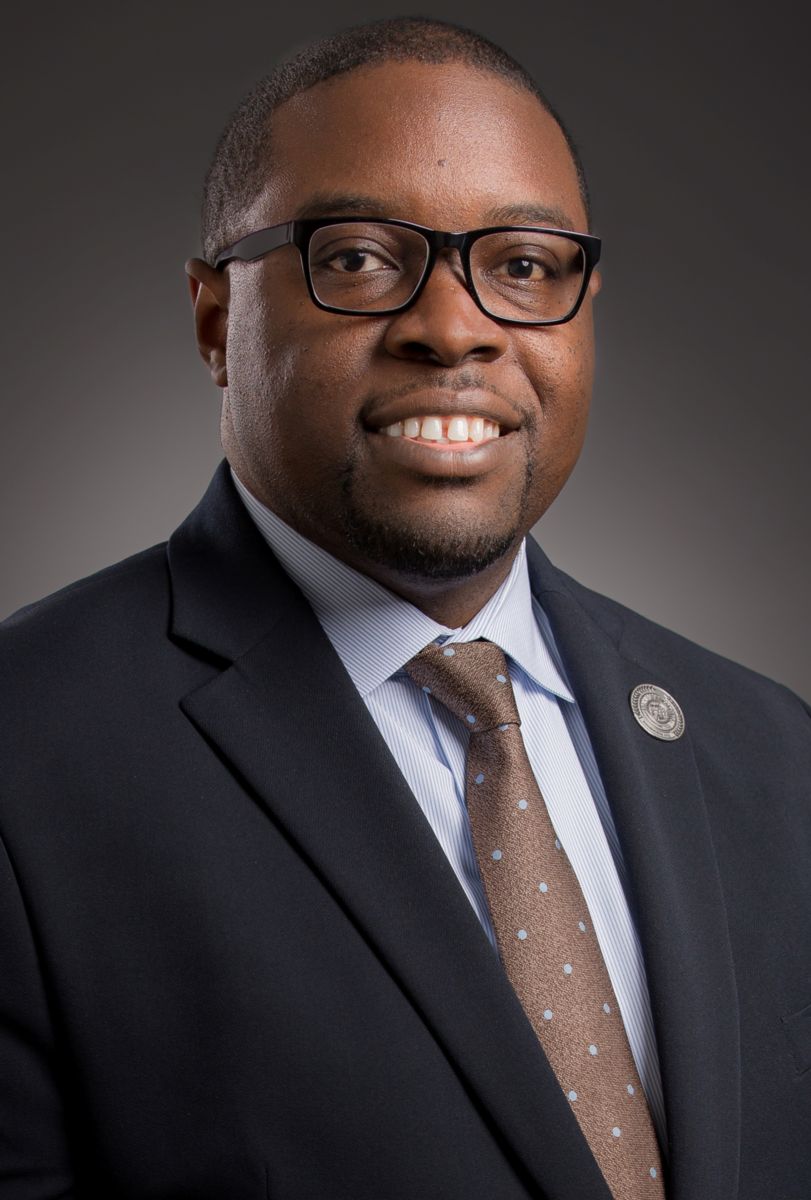Dr. Jermaine Whirl, an African American male wearing a grey suit jacket, a light blue collared shirt, black rimmed glasses, and a cerulean blue tie. A gold pin is on his suit jacket lapel.